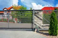 A modern horizontal metal slat gate with an automated opener is set beside a stone pillar, with red-roofed houses and trees in the background.