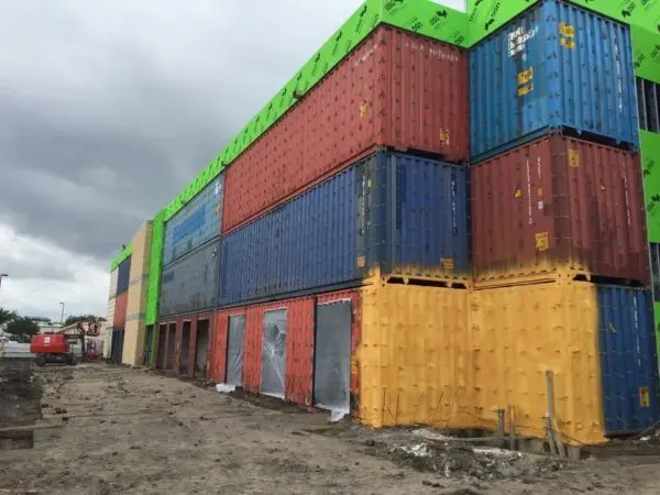 Stacked colorful shipping containers forming a multi-level structure under cloudy skies, with visible construction materials and dirt ground.