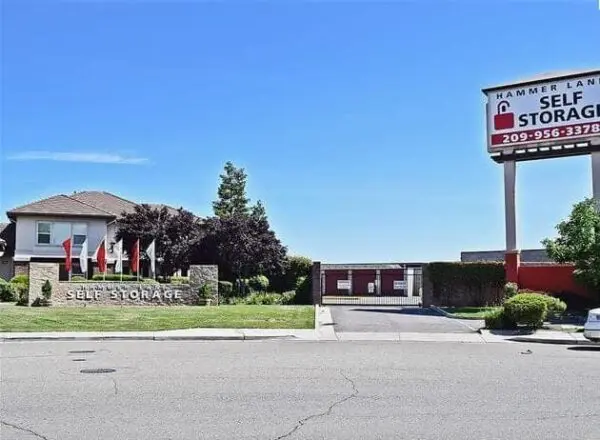 Self storage facility with a gated entrance, surrounded by bushes and flags. A large sign displays contact information. Clear blue sky above.
