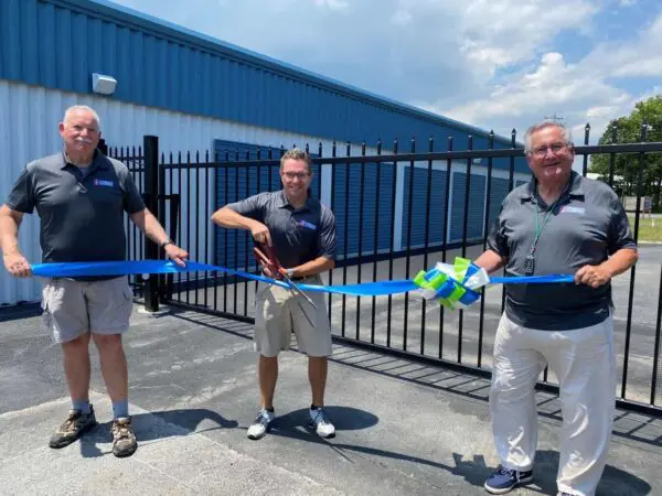 Three men stand outside a building, holding and cutting a blue ribbon with large scissors during a ribbon-cutting ceremony.
