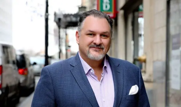 Man with short hair and a beard wearing a blue suit jacket and light shirt, standing on a city street.