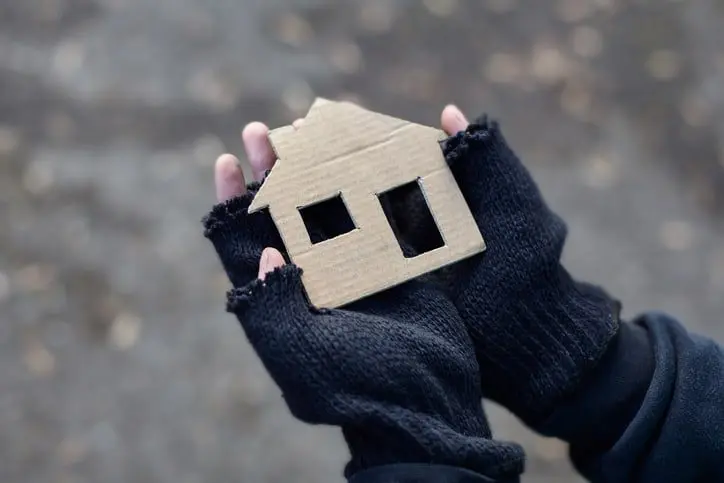 Hands in fingerless gloves holding a cardboard cutout of a house, with a blurred background.