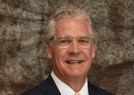 A man with gray hair and glasses, wearing a suit and tie, smiles in front of a textured brown backdrop.