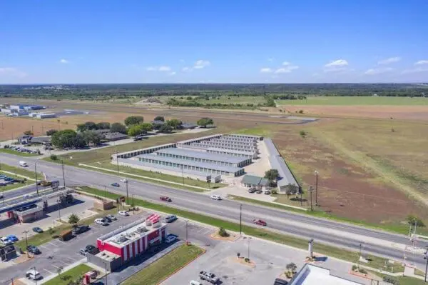 Aerial view of a commercial area including a fast-food restaurant, multiple buildings, a road with cars, and surrounding open fields under a clear blue sky.