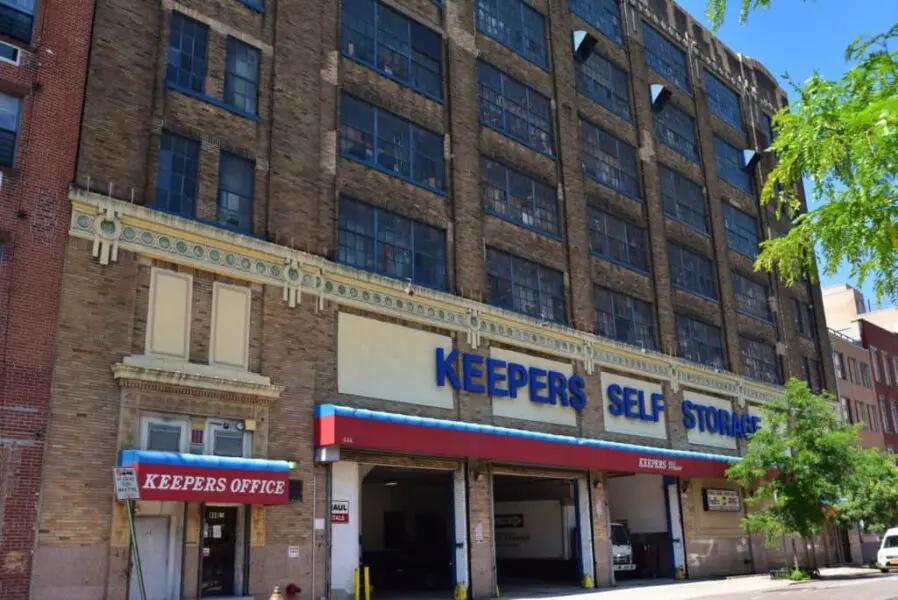 Front view of a multi-story brick building labeled "Keepers Self Storage," with large windows and a sign for "Keepers Office" near the entrance.