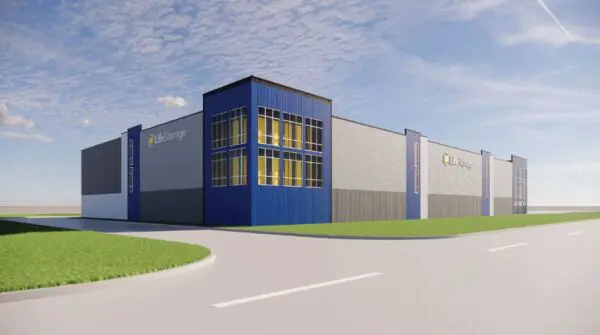 A large, modern storage facility with blue and gray walls is situated beside a road under a partly cloudy sky.