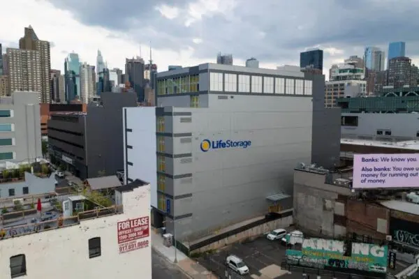 A large Life Storage building in an urban area with a "For Lease" sign on a nearby rooftop. Skyscrapers and cloudy sky in the background.