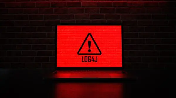 Laptop screen displaying a red warning triangle with an exclamation mark and the text "LOG4J" against a background of binary code, set against a dark brick wall backdrop.
