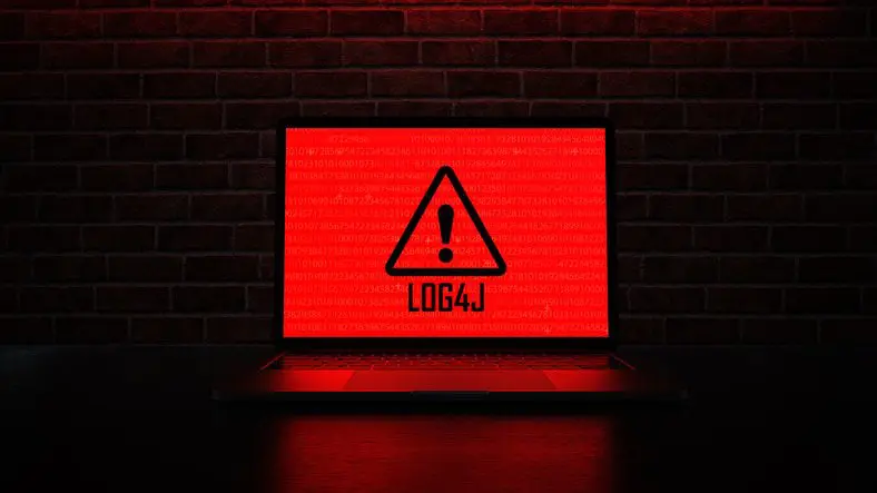 Laptop screen displaying a red warning triangle with an exclamation mark and the text "LOG4J" against a background of binary code, set against a dark brick wall backdrop.
