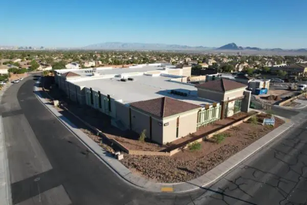 Aerial view of a large, rectangular, beige building with a flat roof, surrounded by a suburban neighborhood and distant mountains in the background.