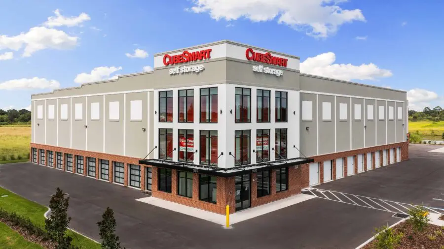 CubeSmart self storage building with large windows, red signage, and multiple garage-style doors. Paved parking lot and a grassy area surround it, under a partly cloudy sky.