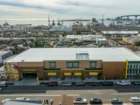 Aerial view of an industrial area with a large building labeled "LOADING ZONE." Cars are parked along the street. Cranes and a bridge are visible in the background.