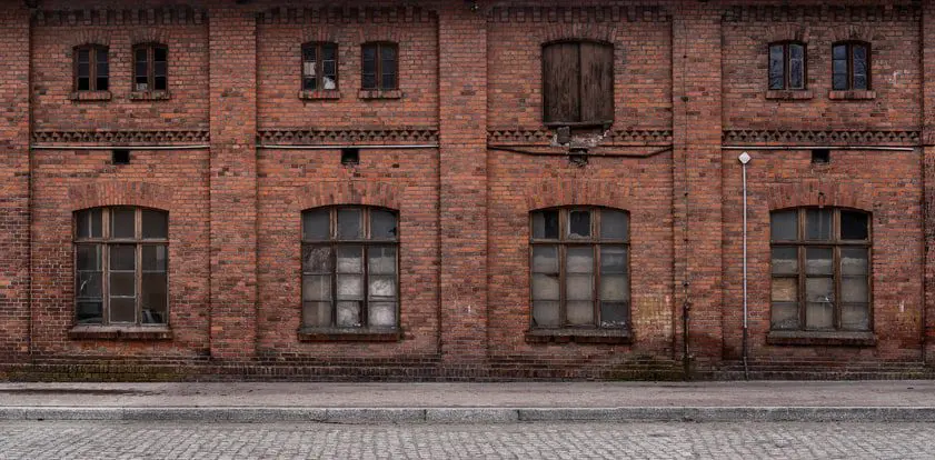 Old brick building with six large windows, some broken or boarded. A wooden double door is above the center window. A cobblestone street runs in front of the building.
