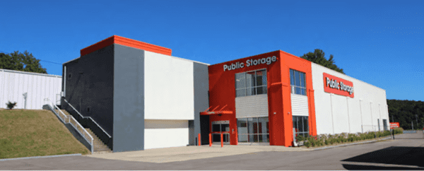 A modern public storage facility with orange and white accents stands under a clear blue sky, featuring a large sign and a few trees nearby.