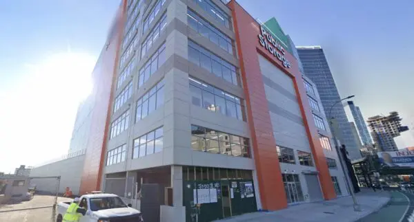 Multi-story storage facility with a bright orange facade, large glass windows, and "Storage" sign. Sunlight shines from the left. Vehicles and a worker are at the entrance. Urban setting.