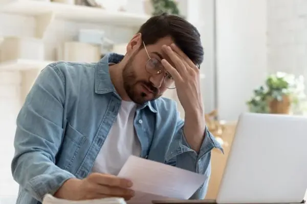 A man in glasses and a denim shirt looks at a document, holding his forehead, seated at a table with a laptop.