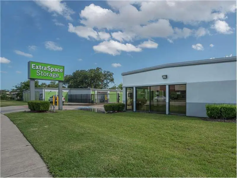 Storage facility with a green sign reading "Extra Space Storage." The building is white with large windows, surrounded by grass and a clear blue sky with clouds.