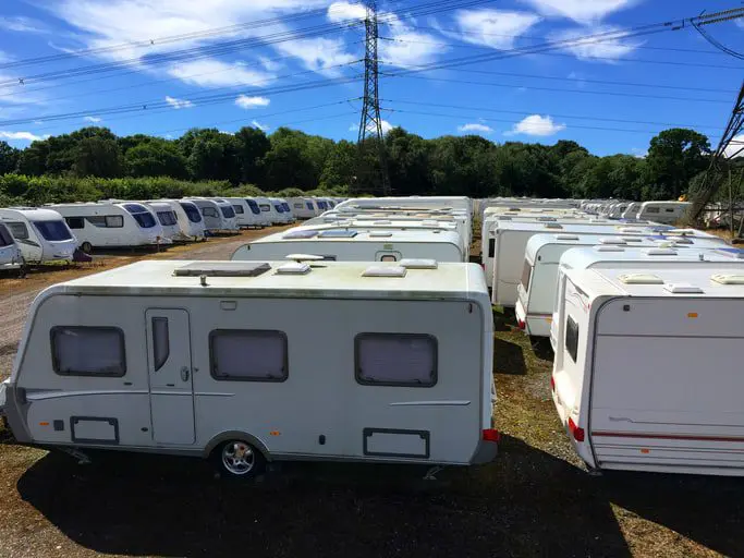 A large number of white caravans are parked in an outdoor storage area under a clear blue sky, with trees in the background and an electricity pylon towering overhead.