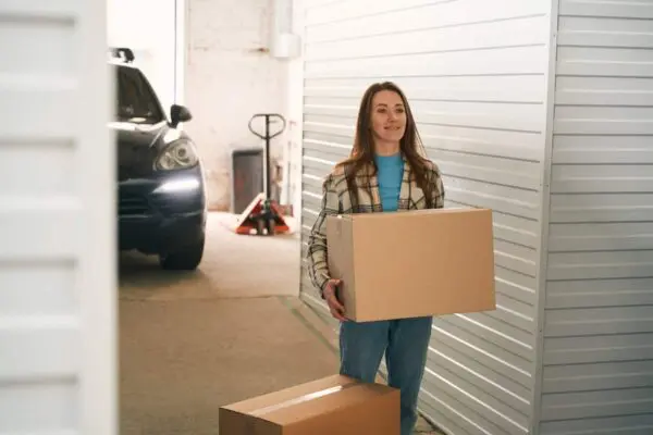 Woman holding a cardboard box stands by a storage unit door. Another box is on the ground. A parked car and a pallet jack are visible in the background.