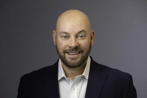 Bald man with a beard wearing a white shirt and dark blazer smiles in front of a plain gray background.