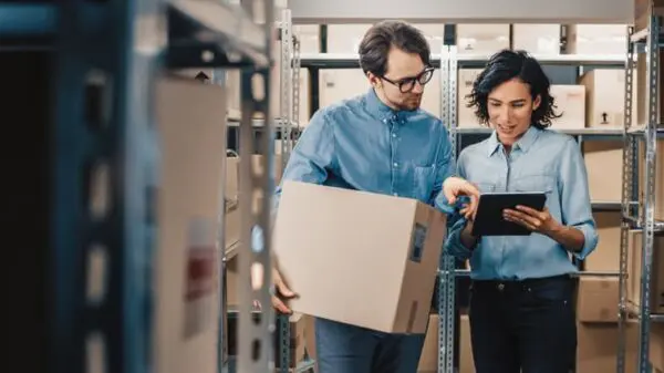 Two people in a warehouse, one holding a box, the other using a tablet, stand among shelves filled with packages.