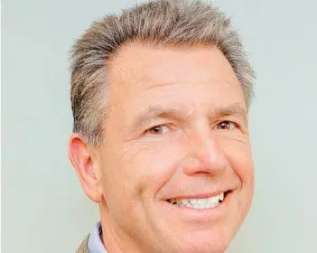Smiling man with short gray hair in a jacket and shirt against a plain background.