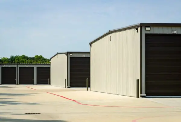 A row of beige storage units with closed brown doors, situated on a concrete lot. The sky is clear and blue, and trees are visible in the background.