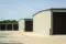 A row of beige storage units with closed brown doors, situated on a concrete lot. The sky is clear and blue, and trees are visible in the background.
