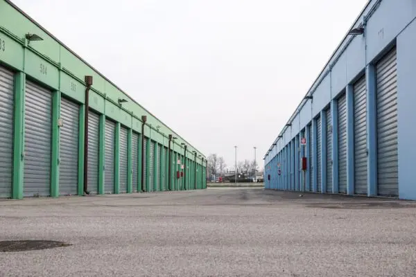 A row of green storage units on the left and blue storage units on the right, with metal roll-up doors, lining a wide concrete path under an overcast sky.