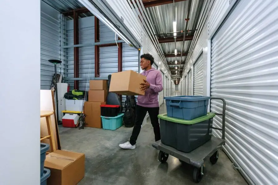 Person carrying a cardboard box down an aisle in a storage facility, with stacked boxes and items nearby.