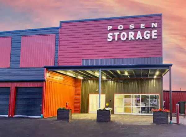 Red and black storage facility with "Posen Storage" sign. Three garage doors and a glass entrance are visible under an overhanging roof. Sky at sunset in the background.