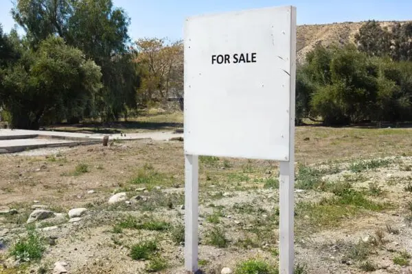 Blank "For Sale" sign in a barren, grassy area with trees and hills in the background.