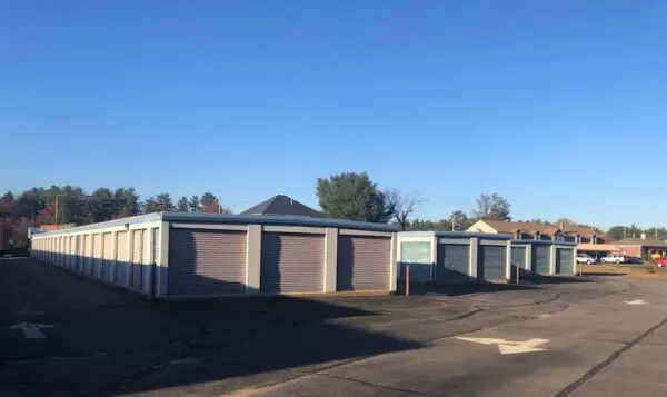 Rows of storage units with closed doors under a clear blue sky, surrounded by a paved lot and trees in the background.
