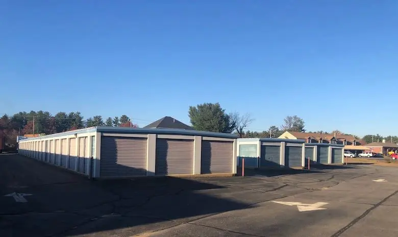 Rows of storage units with closed doors under a clear blue sky, surrounded by a paved lot and trees in the background.