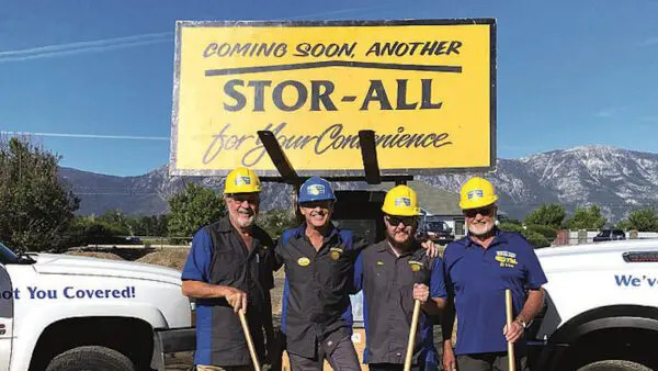Four workers in blue shirts and yellow hard hats stand in front of a "STOR-ALL" sign, holding shovels. Two trucks are parked nearby. Mountains are visible in the background.