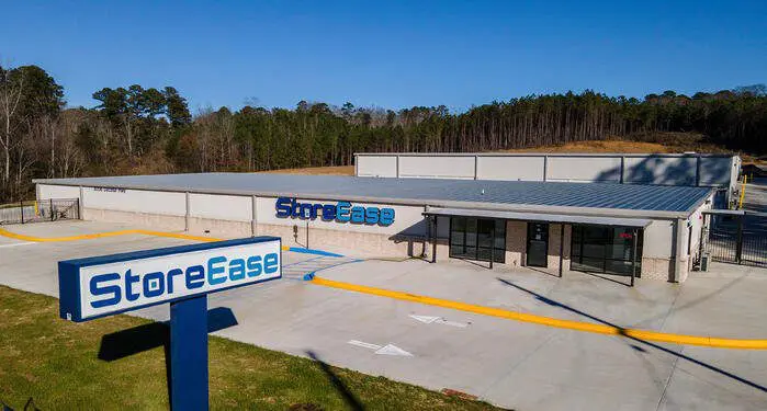 Single-story StoreEase building with a parking lot and a sign in front, surrounded by trees under a clear blue sky.
