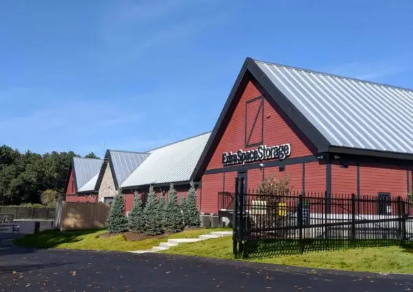 A row of red buildings with gray metal roofs labeled "Extra Space Storage," surrounded by a black fence and small trees, under a clear blue sky.