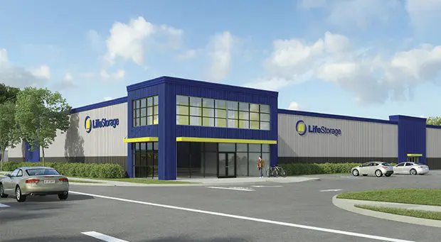 A Life Storage facility with a blue and gray exterior, featuring large windows and entrances, is situated next to a parking area and road with a car and a few bicycles parked outside.