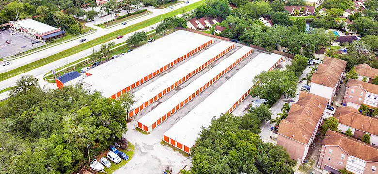 Aerial view of a storage facility with four long buildings surrounded by trees, adjacent to a road and residential area.
