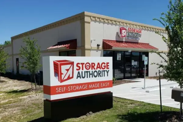 A single-story Storage Authority facility with beige walls and red roofing. A large sign in front reads "SELF-STORAGE MADE EASY." Several young trees are planted around the building.