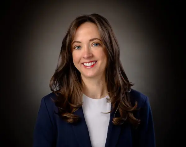 A woman with long brown hair, wearing a dark blazer and white shirt, smiles while posing against a dark gradient background.