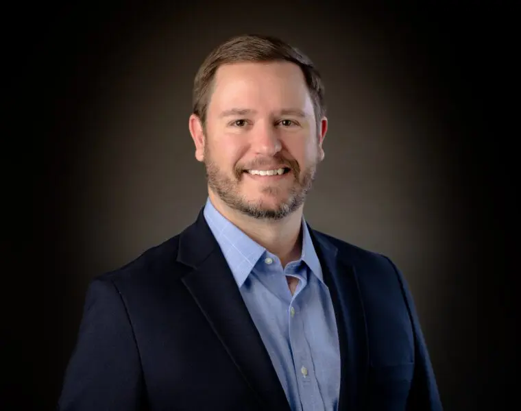 A man with a short beard and neatly combed hair is smiling. He is wearing a dark blazer over a light blue shirt against a plain dark background.