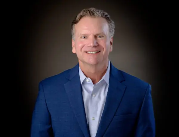 Smiling man with short hair in a blue suit jacket and white shirt, against a dark background.