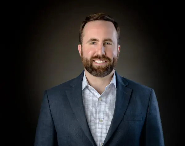 A man with a beard smiles, wearing a blue suit and checkered shirt, against a dark background.