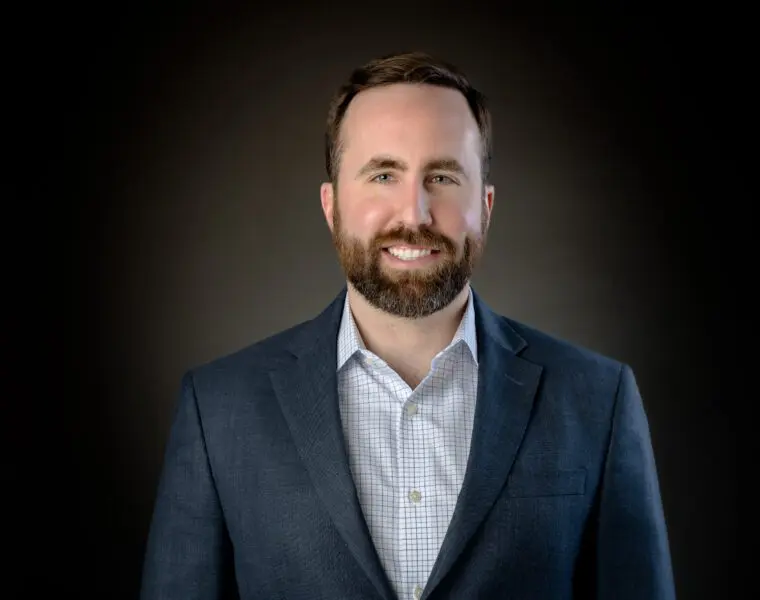 A man with a beard smiles, wearing a blue suit and checkered shirt, against a dark background.