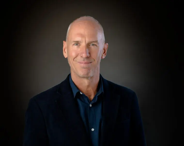 A bald man in a blue shirt and dark blazer smiles slightly against a dark background.