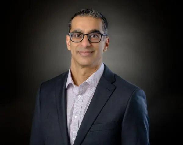 A person wearing glasses and a suit jacket over a collared shirt, posed in front of a dark background.