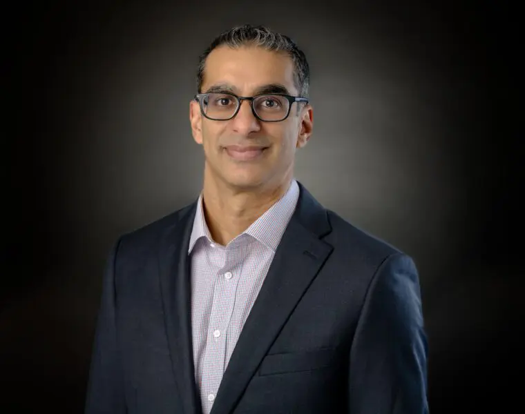 A person wearing glasses and a suit jacket over a collared shirt, posed in front of a dark background.