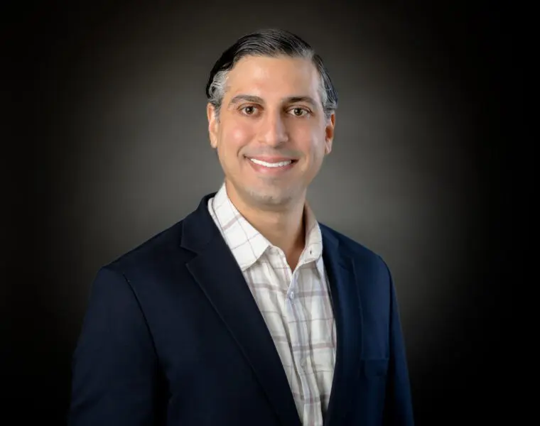 A person with short dark hair wearing a plaid shirt and a dark blazer smiles at the camera against a dark background.
