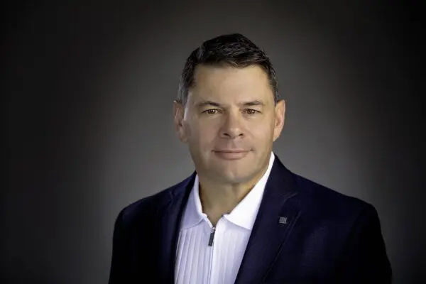 Man in a dark suit and white shirt poses against a neutral background, looking directly at the camera.